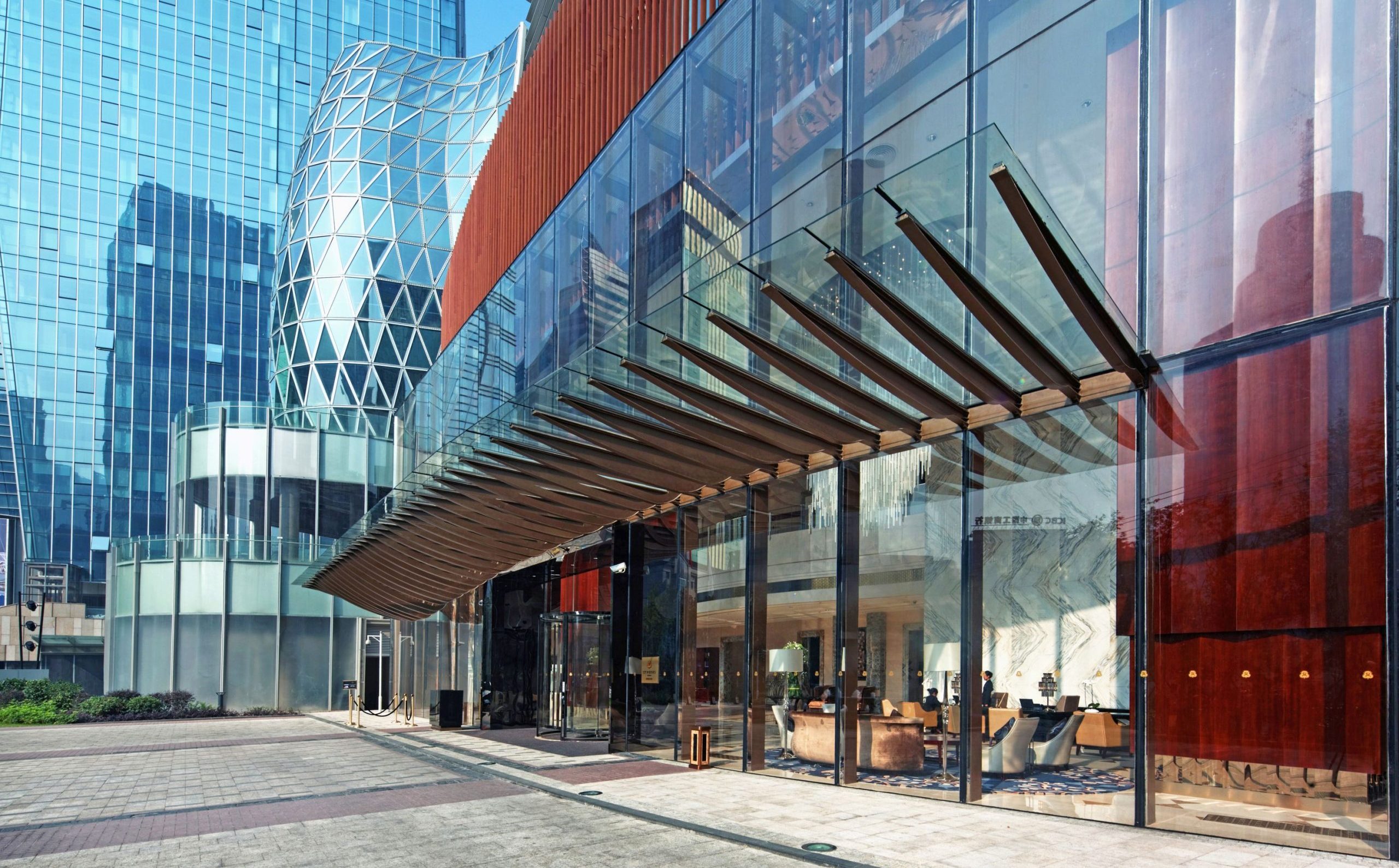 Aedas’ Evergrande Hua Zhi Plaza with glass hotel entrance canopy, vertical fins and mixed‑use towers forming an urban oasis. Street‑level view of Aedas‑designed Evergrande Hua Zhi Plaza showing a transparent glass entrance canopy with cantilevered steel fins, vertical terracotta‑toned façade fins, and high‑performance glazed towers above. The ground plane integrates retail frontage and sheltered public circulation, linking hotel, office and commercial components within a landscaped mixed‑use urban complex.