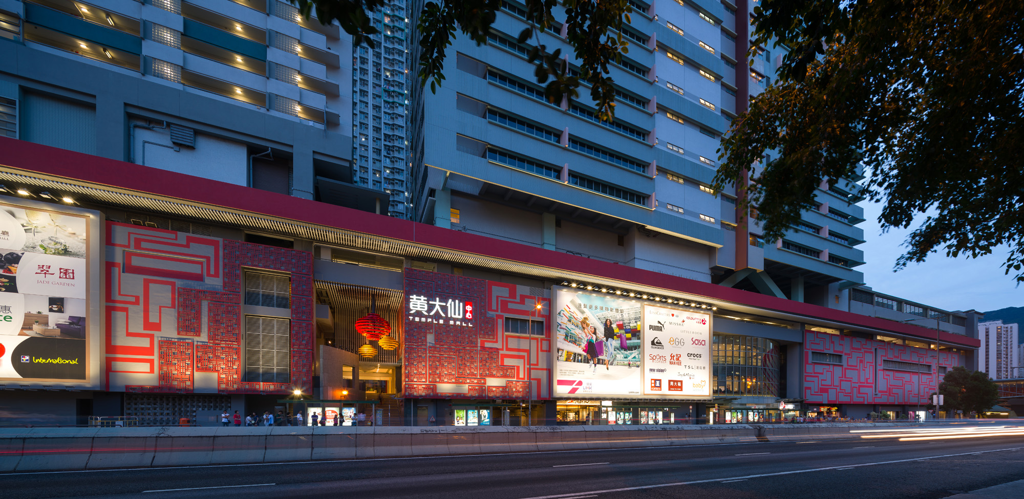 Aedas’ Temple Mall in Hong Kong is shown at night as a revitalised neighbourhood shopping centre with an illuminated red patterned façade, large digital billboards, and continuous retail frontage beneath residential towers. The image highlights a contemporary architectural upgrade that enhances street presence, improves wayfinding, and reinforces Temple Mall’s role as a vibrant community retail hub within Wong Tai Sin.