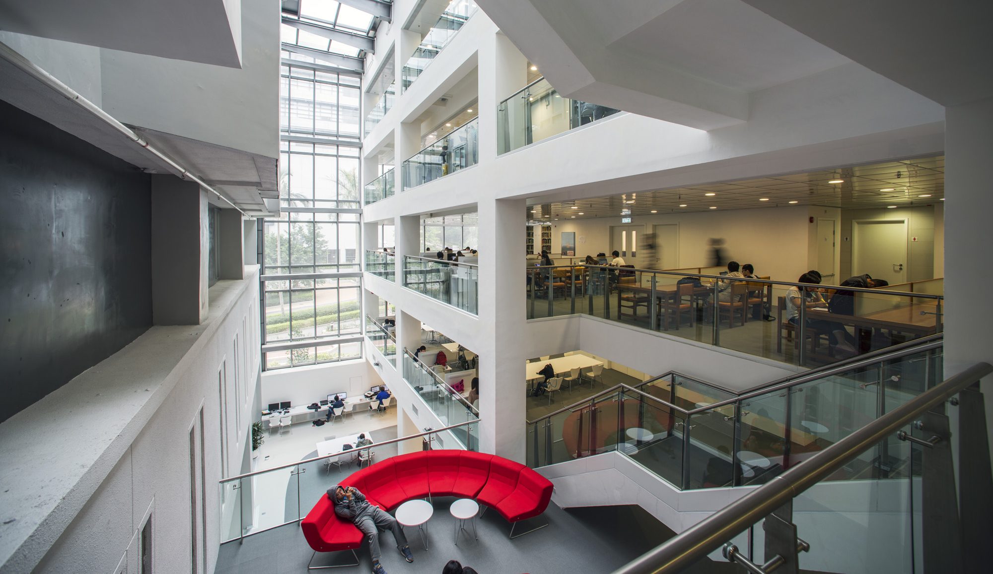 Aedas’s CUHK Library Extension interior is shown as a tall, multi‑level atrium with stacked study floors, glass balustrades, exposed concrete structure, and a full‑height glazed skylight. The image highlights sectional clarity, daylight‑driven academic space, and vertically integrated circulation within a university library building.