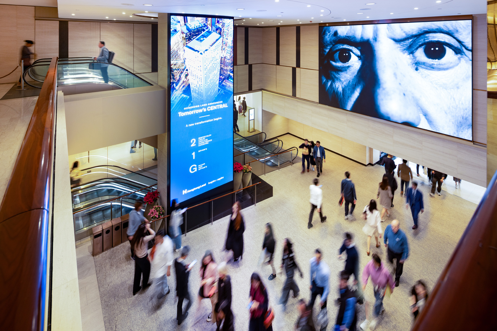 Aedas’ Chater House in Hong Kong is shown as a lively, multi‑storey retail atrium with polished stone floors, warm timber balustrades, and large‑scale digital media walls. The image highlights a reconfigured Central District interior where escalators, generous circulation, and luxury storefronts—including a flagship fashion space—create an active urban shopping environment integrated seamlessly beneath a Grade‑A office tower.