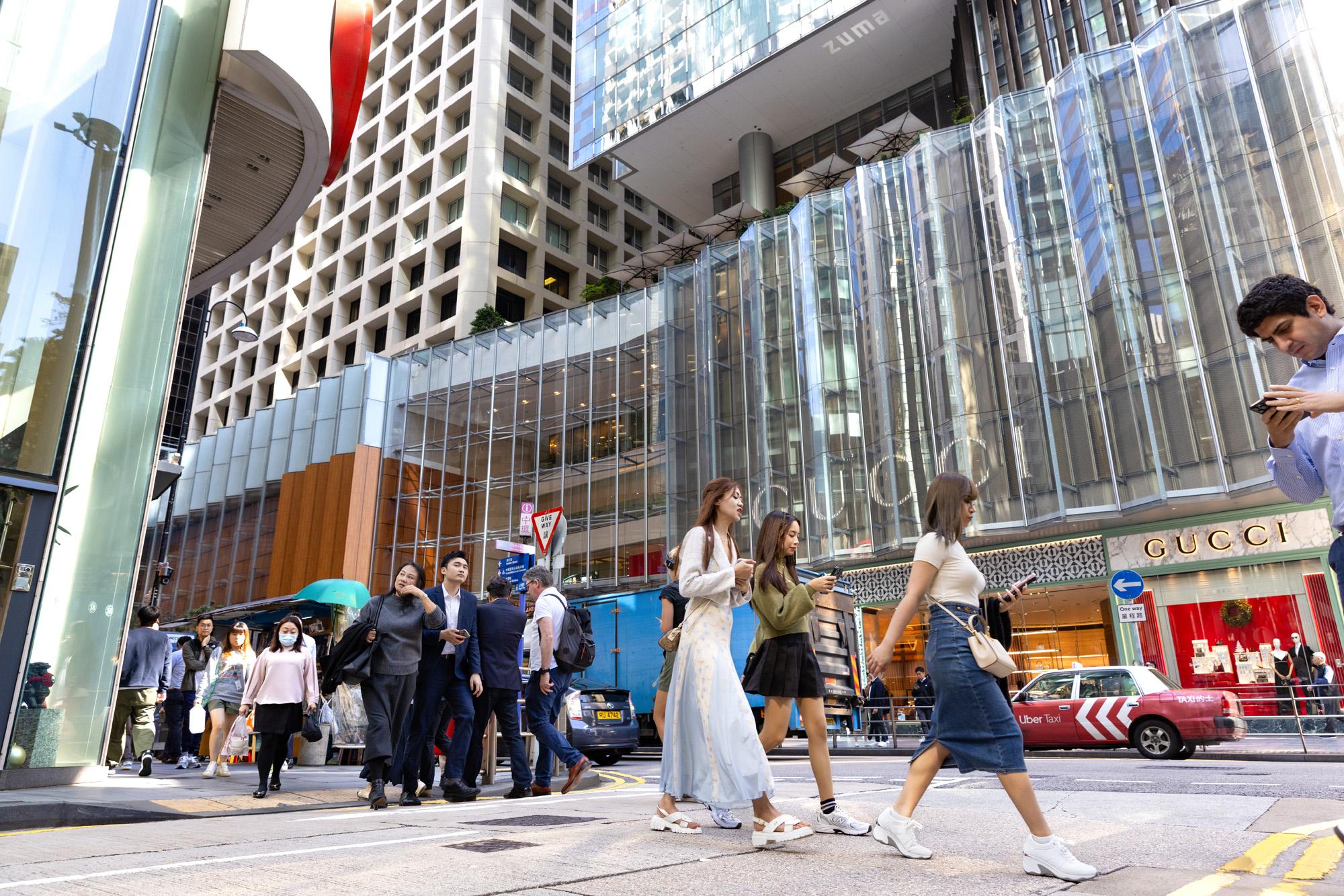 Aedas’ Landmark in Hong Kong is shown as a revitalised luxury retail and office complex with transparent glass façades, vertically layered atrium volumes, and prominent two‑storey flagship storefronts. The image highlights an active Central streetscape where high‑end retail, digital façades, and refined architectural detailing animate pedestrian movement beneath the rebuilt Landmark East tower and integrated hotel and mall precinct.