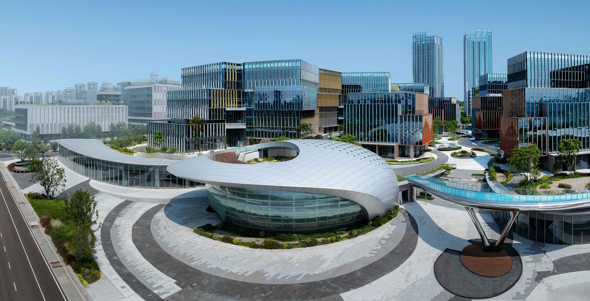 Aedas’ Chengdu Intelligent Harbour - campus‑scale mixed‑use development with oval pavilion, glass‑clad offices and landscaped pedestrian network. Aerial view of an Aedas‑designed mixed‑use campus featuring an oval low‑rise pavilion with curved roof and continuous glazing, surrounded by mid‑rise office blocks with unitised glass façades and vertical fins. The site includes integrated pedestrian loops, elevated walkways, and extensive landscape planting within a high‑density urban district.