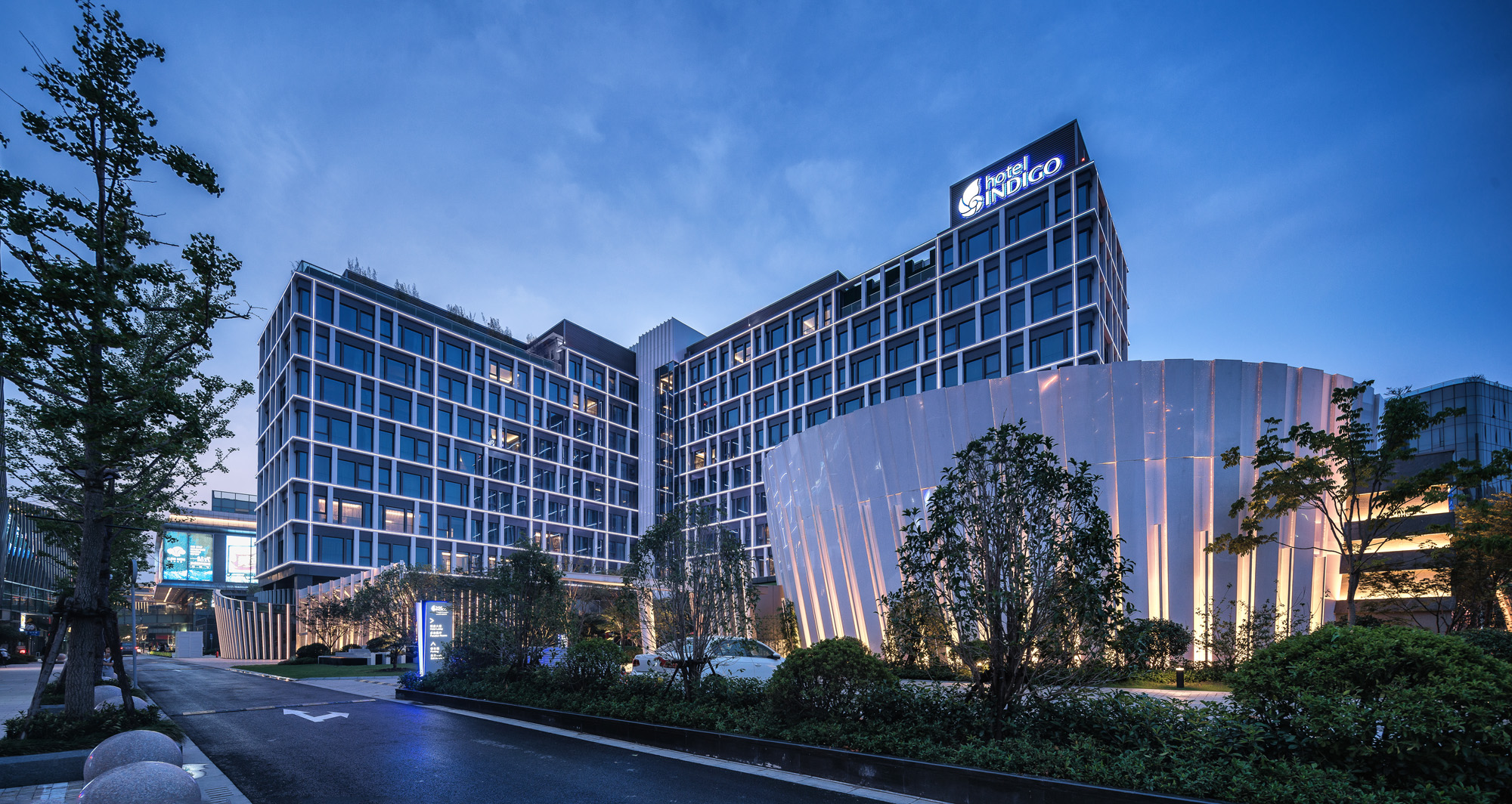 Aedas’ Hotel Indigo at Hongqiao Paradise Walk in Shanghai is shown as a distinctive hotel complex with a curvilinear metallic podium resembling a textured shell, set beneath a rectilinear guestroom tower. The image highlights a dramatic arrival sequence with landscaped forecourts, layered façade rhythms, and illuminated glazing that establishes the hotel as a prominent architectural landmark within the mixed‑use retail district.
