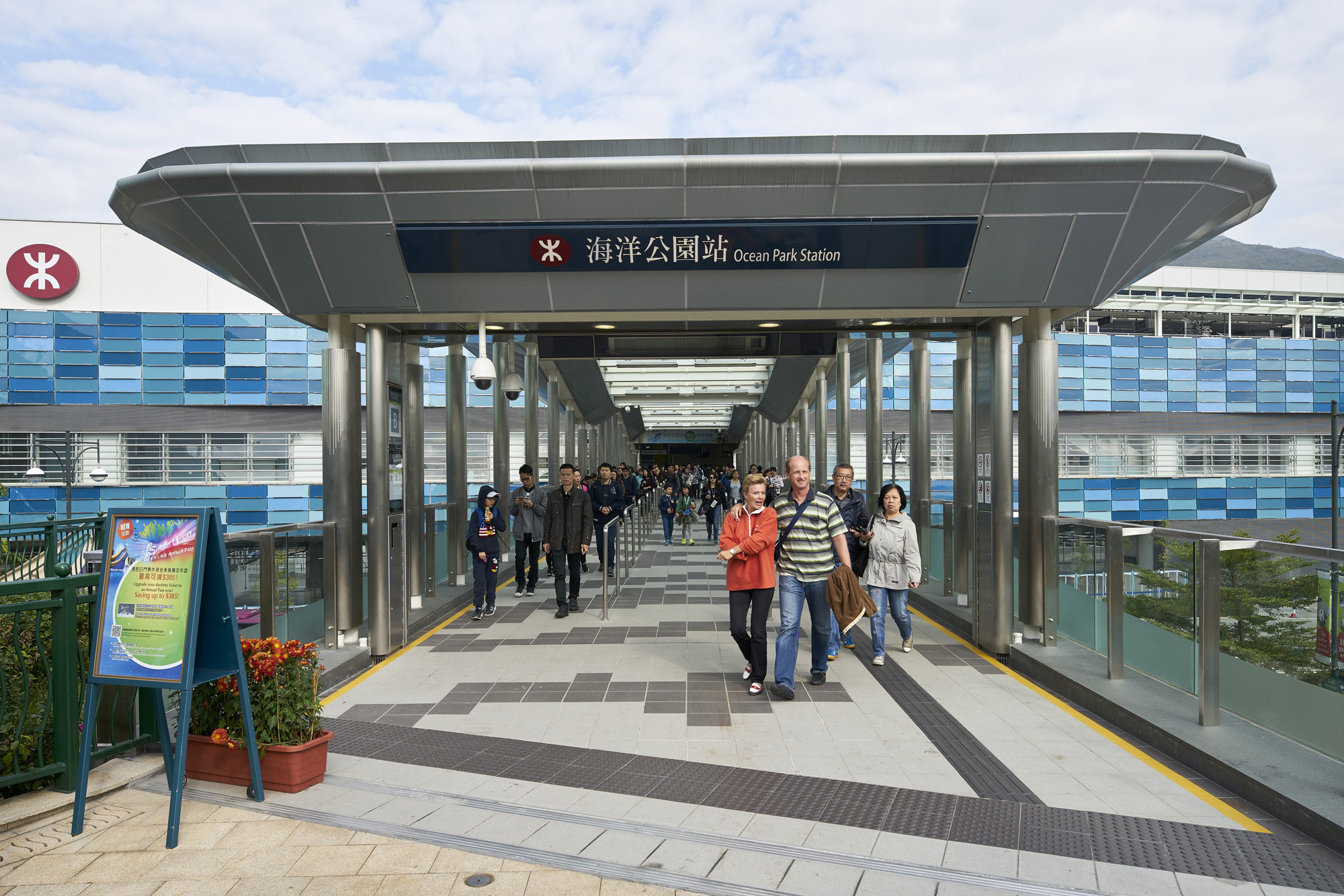 Aedas’s Ocean Park Station on the MTR South Island Line is shown with an elevated pedestrian approach, steel column supports, long-span canopy roof, glazed balustrades, and integrated station signage. The image highlights transport architecture focused on weather protection, passenger flow, and seamless connection between station and surrounding urban landscape in Hong Kong.