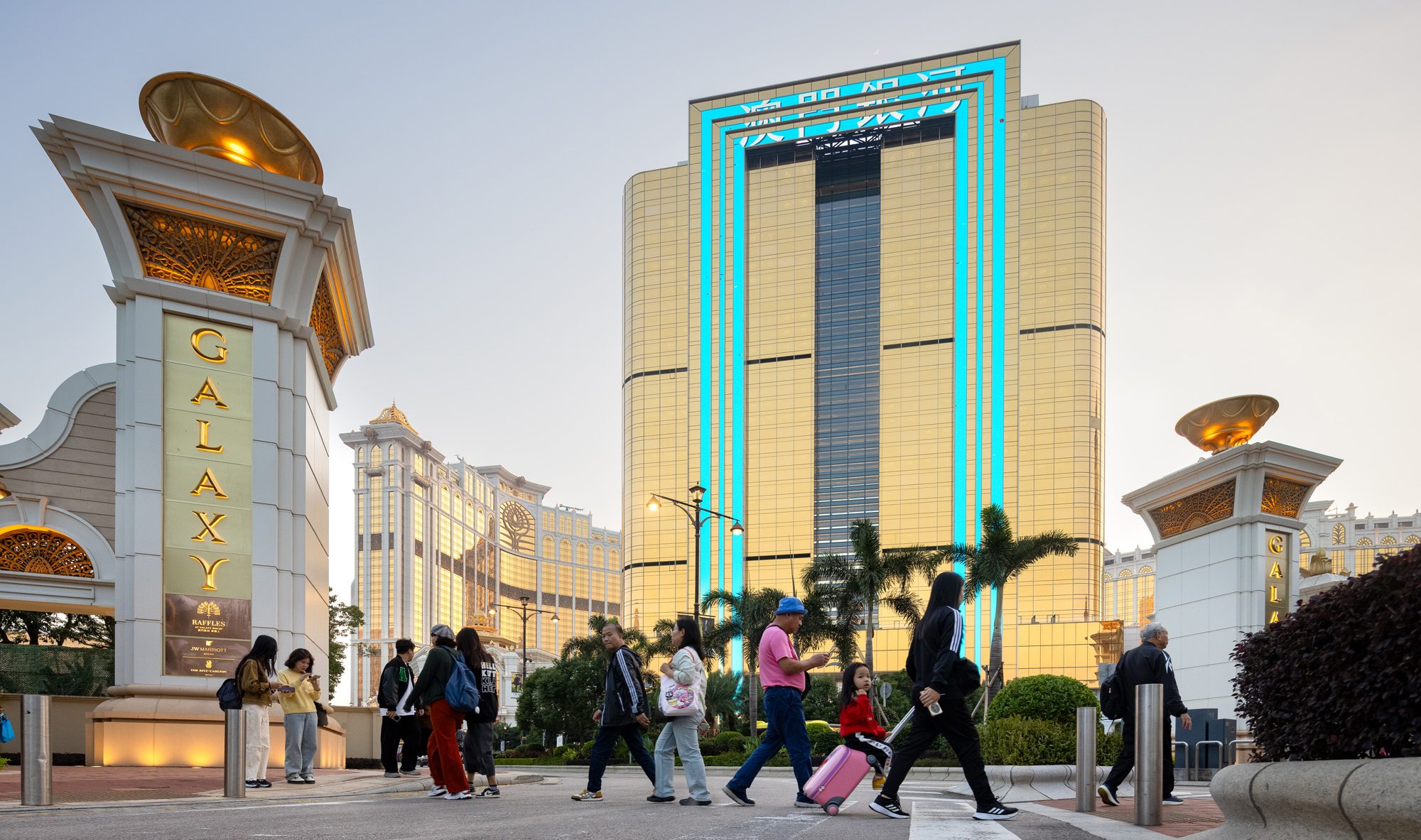 Aedas’ Raffles at Galaxy Macau with gold‑toned glazed hotel tower, palm‑lined boulevard and resort podium. Street‑level view of Aedas‑designed Raffles at Galaxy Macau showing a tall hotel tower clad in gold‑finished curtain wall glazing, articulated by vertical bands and rounded corners. A landscaped podium and palm‑lined pedestrian crossings front the building, with adjacent Galaxy Macau resort structures defining a high‑density hospitality precinct in Macau’s subtropical climate.