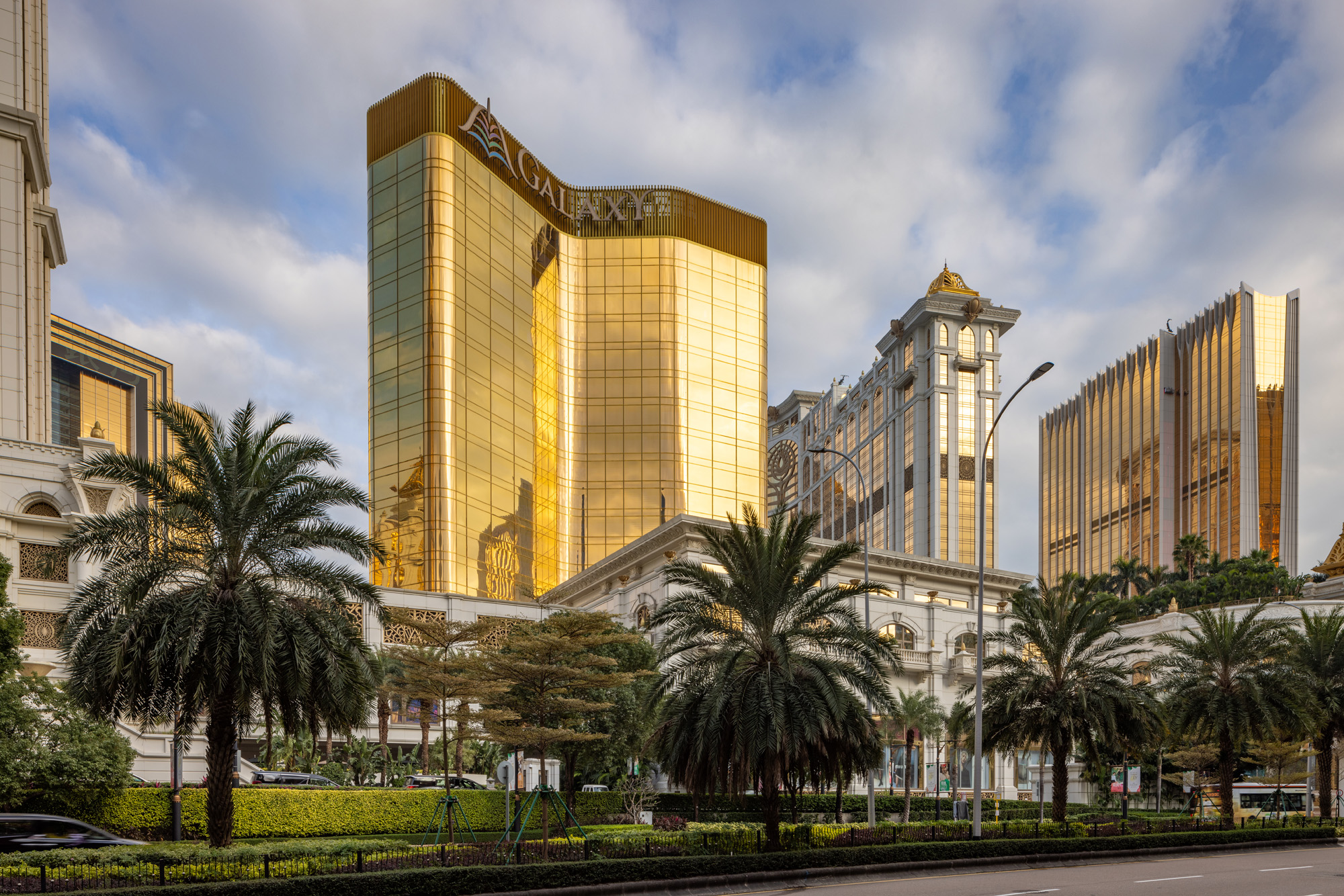 Aedas’ Capella at Galaxy Macau with gold‑toned glazed towers, podium gardens and palm‑lined boulevard in Cotai. Street‑level view of Aedas‑designed Capella at Galaxy Macau showing mid‑rise hotel towers clad in gold‑finished curtain wall glazing with rounded corners above a landscaped podium. Palm‑lined boulevards, layered podium façades and integrated drop‑off zones define the resort frontage within Macau’s high‑density Cotai hospitality district.