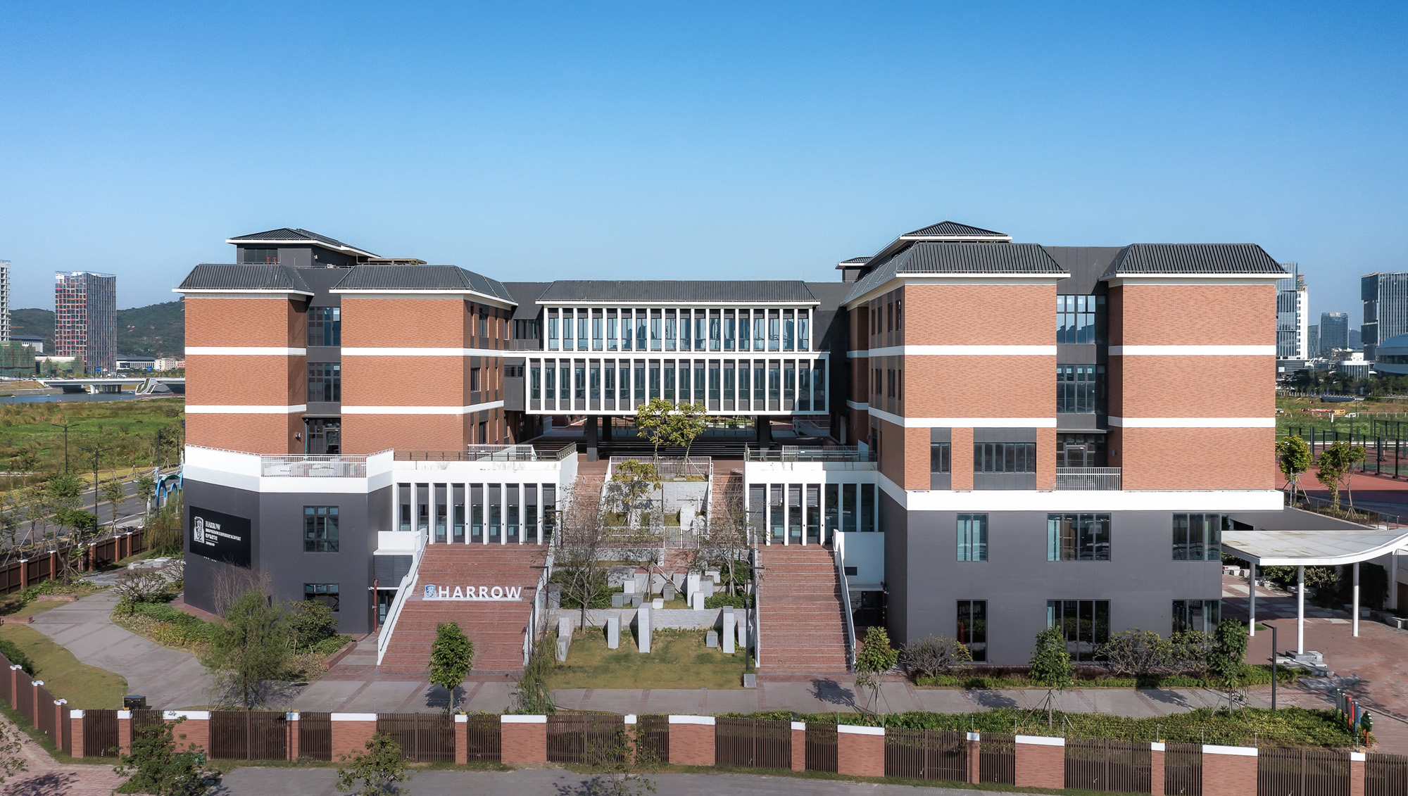 Aedas’ Harrow Innovation Leadership Academy Hengqin with brick‑clad academic blocks and central stepped campus. Aerial frontal view of the Aedas‑designed Harrow Innovation Leadership Academy Hengqin, showing symmetrical academic buildings arranged around a central stepped courtyard. The campus features brick‑clad façades, pitched metal roofs, and deep window recesses with vertical glazing. Terraced outdoor circulation, landscaped lawns, and stair‑linked platforms structure movement across the multi‑level educational complex.