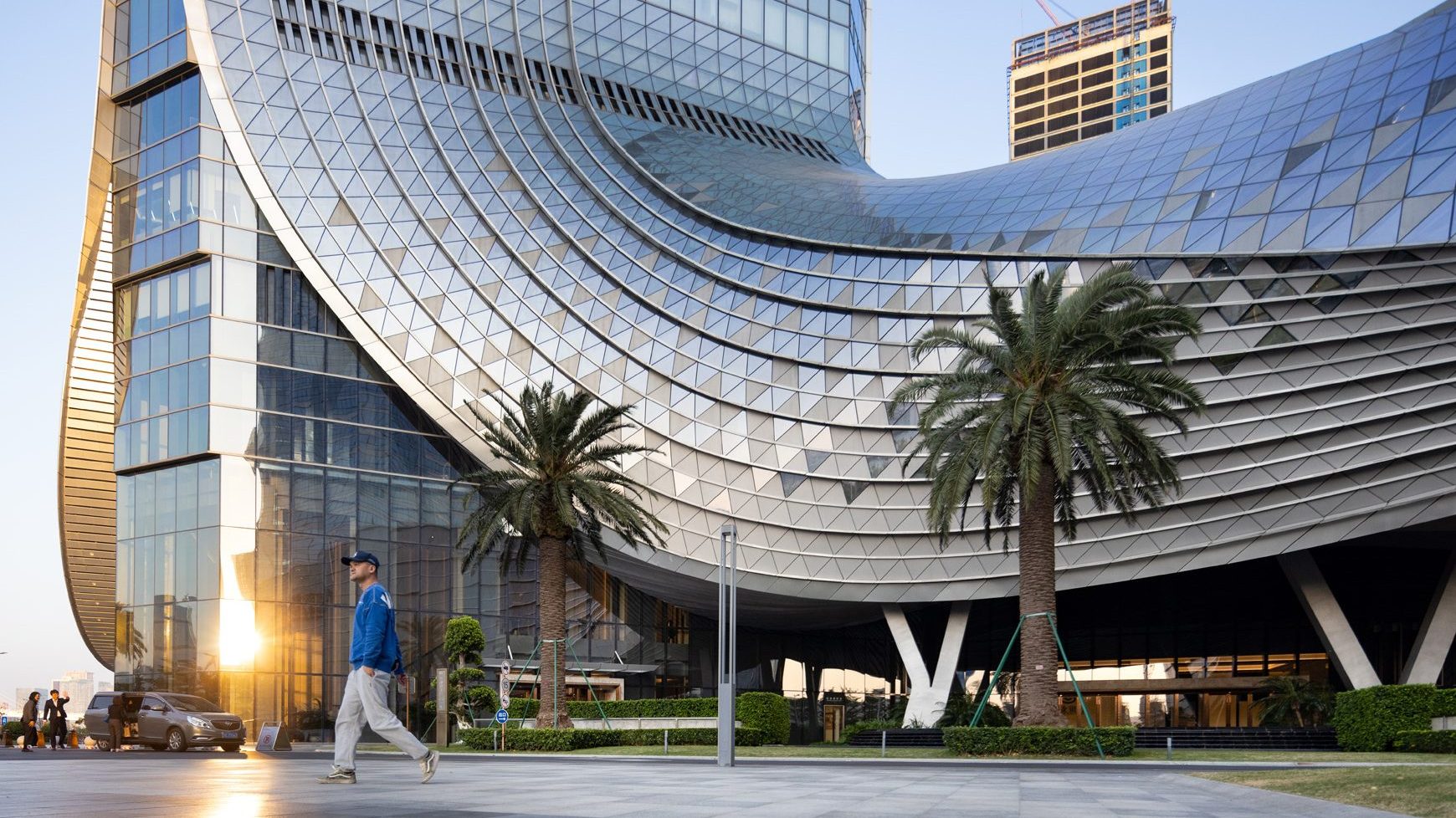 Aedas’ Nansha IFC with curved glass towers, sky garden connection and wave‑inspired façade overlooking the waterfront. Street‑level waterfront view of Aedas‑designed Nansha IFC in Guangzhou, showing two tall office towers with curved, slanted glass curtain walls rising from a sculpted podium. The façades feature undulating horizontal patterns and tilted glazing panels, while a sky garden links the twin towers above, framing landscaped forecourts and coastal public space below.
