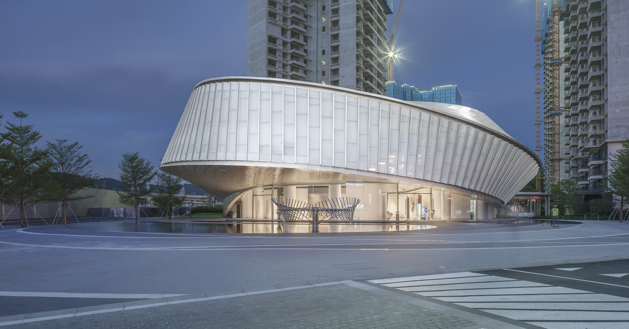 Aedas’ Humen Poly Exhibition Center with curved glazed curtain wall, vertical fins and cantilevered elliptical volume. Dusk exterior of an Aedas-designed TOD gateway pavilion at Humen Poly, formed as a cantilevered elliptical ring over a transparent lobby. The envelope uses vertically folded curtain-wall glazing with dense mullion/fins and a recessed soffit. The forecourt is an open plaza with traffic markings and perimeter planting.