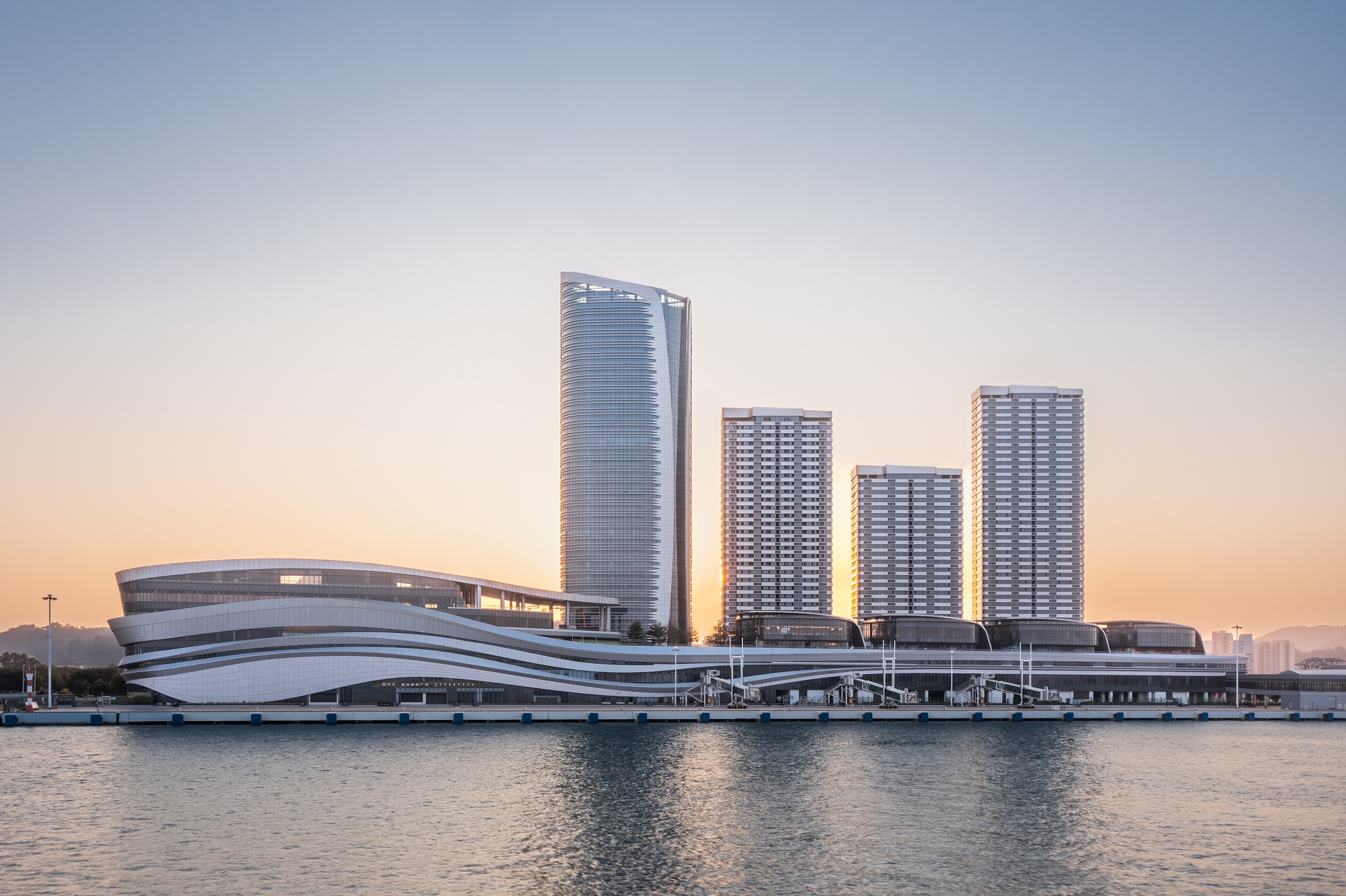 Aedas’ Guangzhou Nansha International Cruise Terminal with whaleboat‑inspired roof and layered waterfront terminal massing. Waterfront view of the Aedas‑planned Guangzhou Nansha International Cruise Terminal, showing a low‑rise terminal with flowing, layered rooflines and horizontal banded façades. The building stretches along the quay with integrated cruise berths and gangway connections, set against high‑rise towers in a coastal delta environment under subtropical climatic conditions.