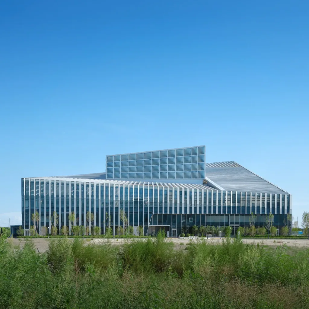 Aedas’ National Dairy Technology Innovation Center in Yili, Hohhot - low‑rise institutional building with louvred glass façade and deep roof canopy in a landscaped campus. Frontal view of an Aedas-designed low-rise institutional building with a rectilinear massing, vertical louvres, and floor-to-floor glazed façade. A stepped roof form and extended canopy provide solar shading. The building sits within an open landscaped site, with planting buffers and setback circulation in a low-density, warm-climate context.