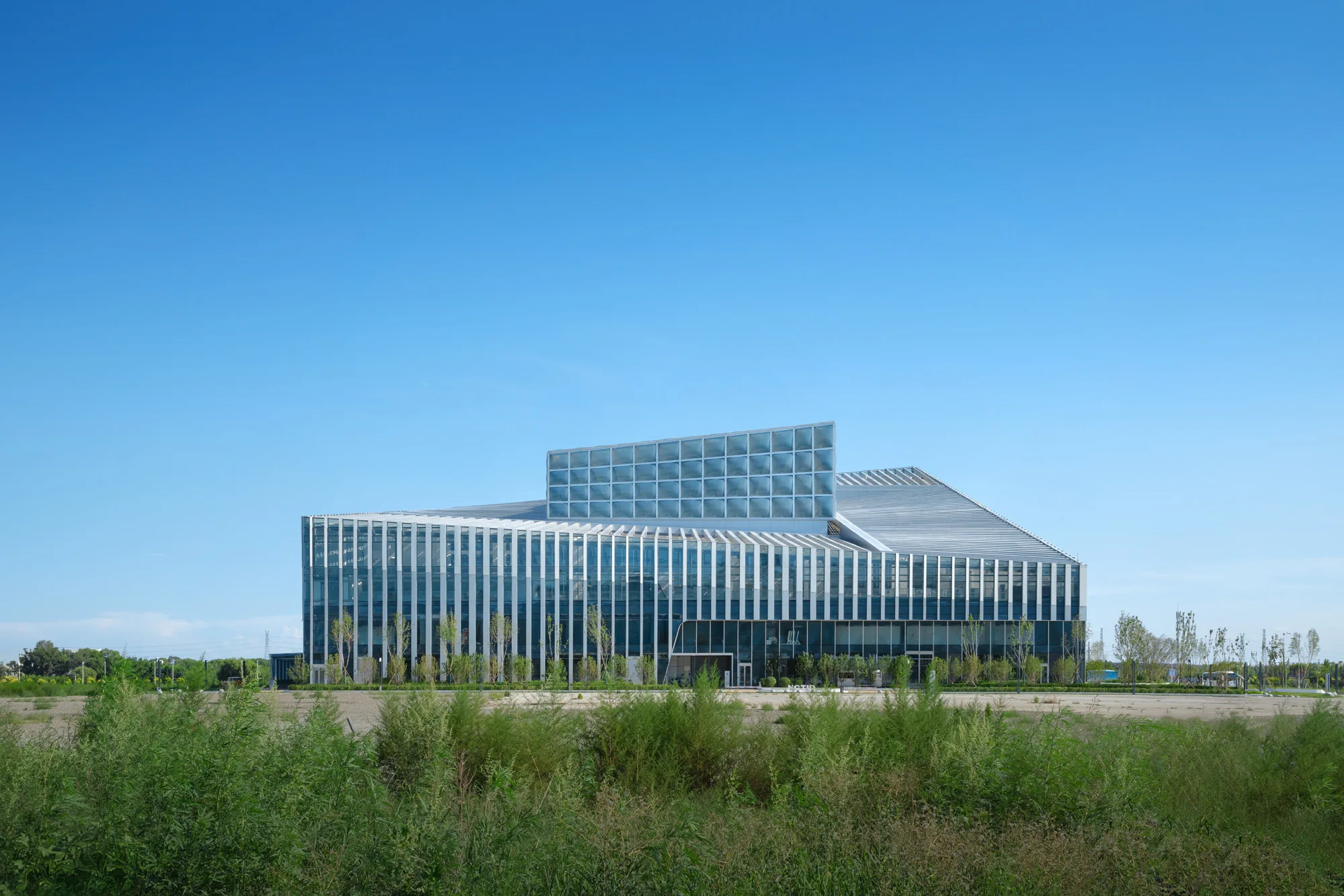 Aedas’ National Dairy Technology Innovation Center in Yili, Hohhot - low‑rise institutional building with louvred glass façade and deep roof canopy in a landscaped campus. Frontal view of an Aedas-designed low-rise institutional building with a rectilinear massing, vertical louvres, and floor-to-floor glazed façade. A stepped roof form and extended canopy provide solar shading. The building sits within an open landscaped site, with planting buffers and setback circulation in a low-density, warm-climate context.