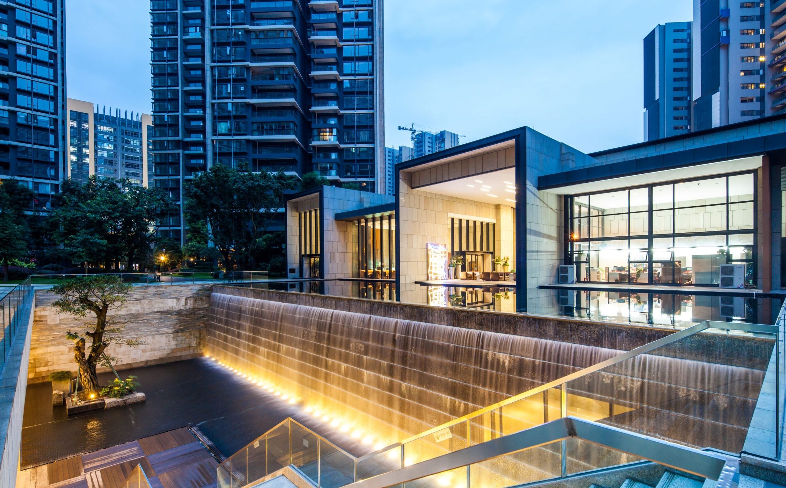 Aedas’ high‑rise residential development with cascading podium, water feature and landscaped central garden court. Evening courtyard view of an Aedas‑designed high‑density residential development, showing multiple residential towers arranged around a terraced podium landscape. A stepped water feature spills through cascading car‑park levels, flanked by a clubhouse pavilion with floor‑to‑ceiling glazing. Integrated planting, reflective pools and pedestrian bridges organise circulation within a compact urban block.