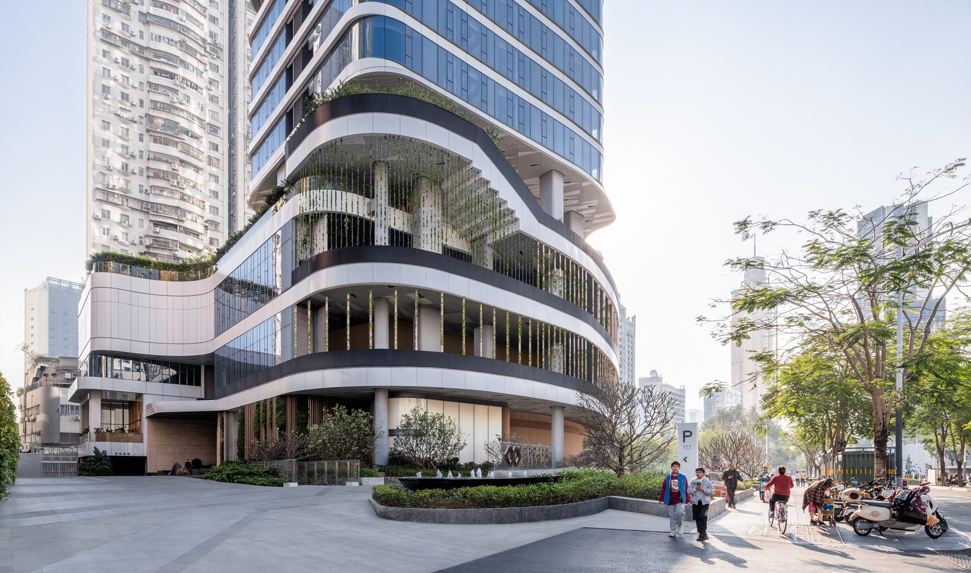Aedas’ Shenzhen Yanlord Luohu Mixed‑use Development is shown at podium level as a sculpted, multi‑storey base with flowing horizontal floor plates, recessed terraces, and vertical greenery integrated into glazed façades. The image highlights a people‑focused residential landmark where curved architectural edges, landscaped public space, and transparent ground‑floor zones create an inviting urban interface beneath the rotated residential tower.