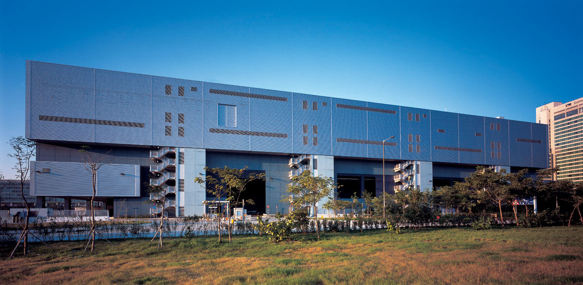 Aedas’s Tradeport Logistics Centre at Hong Kong International Airport is shown as a large‑scale logistics building with a long rectilinear form, solid industrial façade, wide service openings, and elevated undercroft. The exterior highlights airport‑oriented logistics architecture, structural clarity, and high‑throughput industrial massing in a constrained aviation precinct.