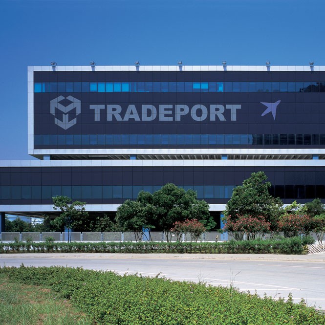Aedas’s Tradeport Logistics Centre at Hong Kong International Airport is shown as a large‑scale logistics building with a long rectilinear form, solid industrial façade, wide service openings, and elevated undercroft. The exterior highlights airport‑oriented logistics architecture, structural clarity, and high‑throughput industrial massing in a constrained aviation precinct.