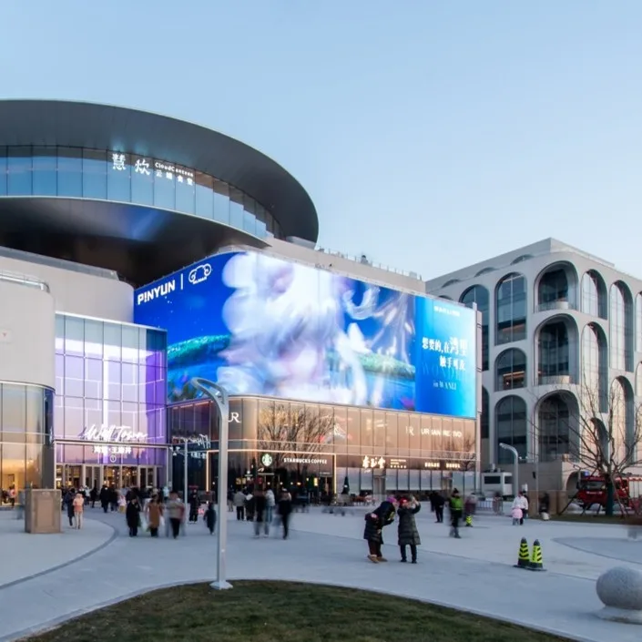Aedas’s Well Town and NOUS Land in Beijing is shown as a large mixed‑use destination with stepped retail volumes, curved podium forms, integrated digital media façades, and expansive pedestrian plazas. The street‑level view highlights inward‑focused commercial circulation, terraced green edges, and direct connection to surrounding transport and cultural infrastructure.
