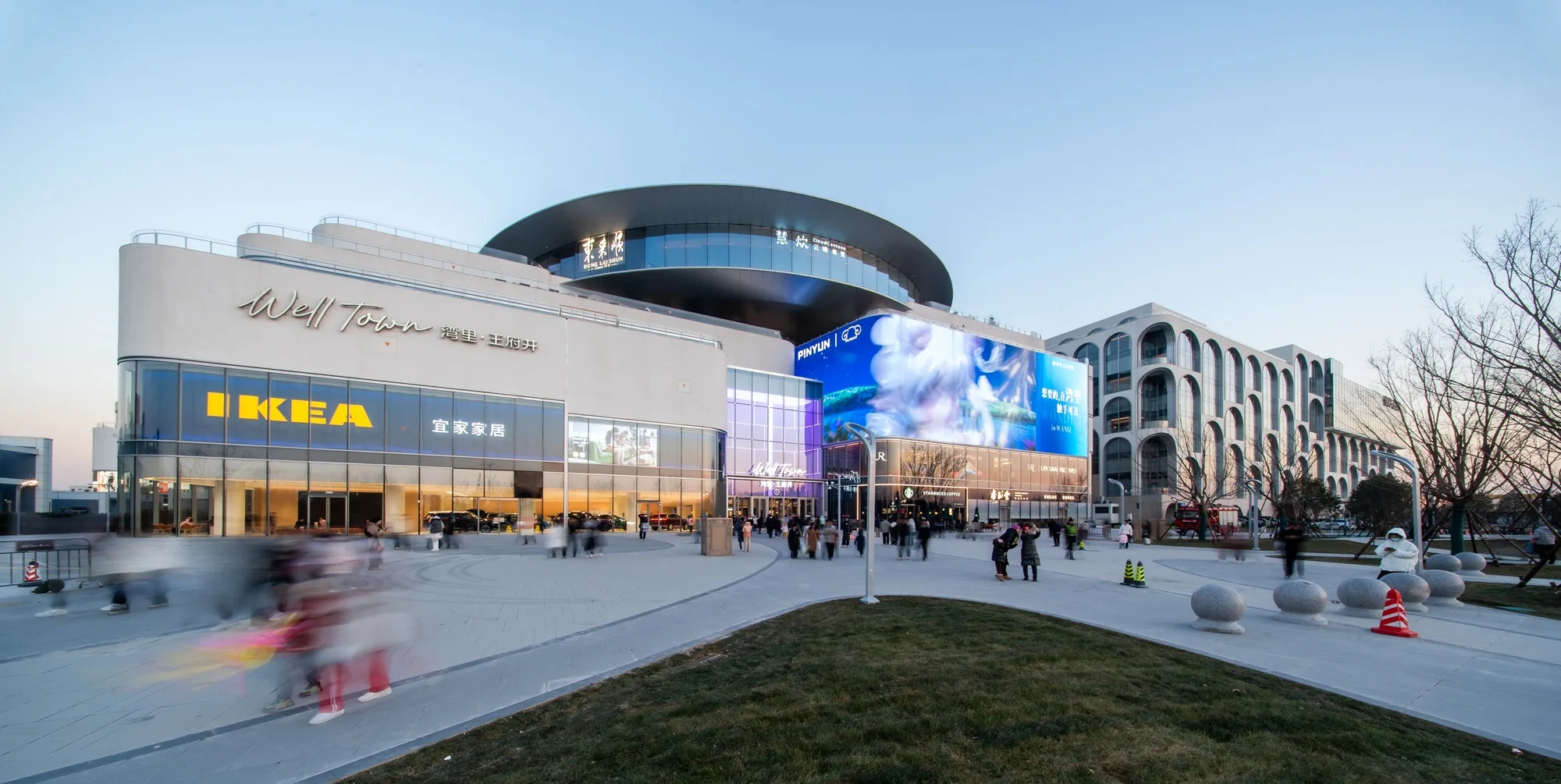 Aedas’s Well Town and NOUS Land in Beijing is shown as a large mixed‑use destination with stepped retail volumes, curved podium forms, integrated digital media façades, and expansive pedestrian plazas. The street‑level view highlights inward‑focused commercial circulation, terraced green edges, and direct connection to surrounding transport and cultural infrastructure.