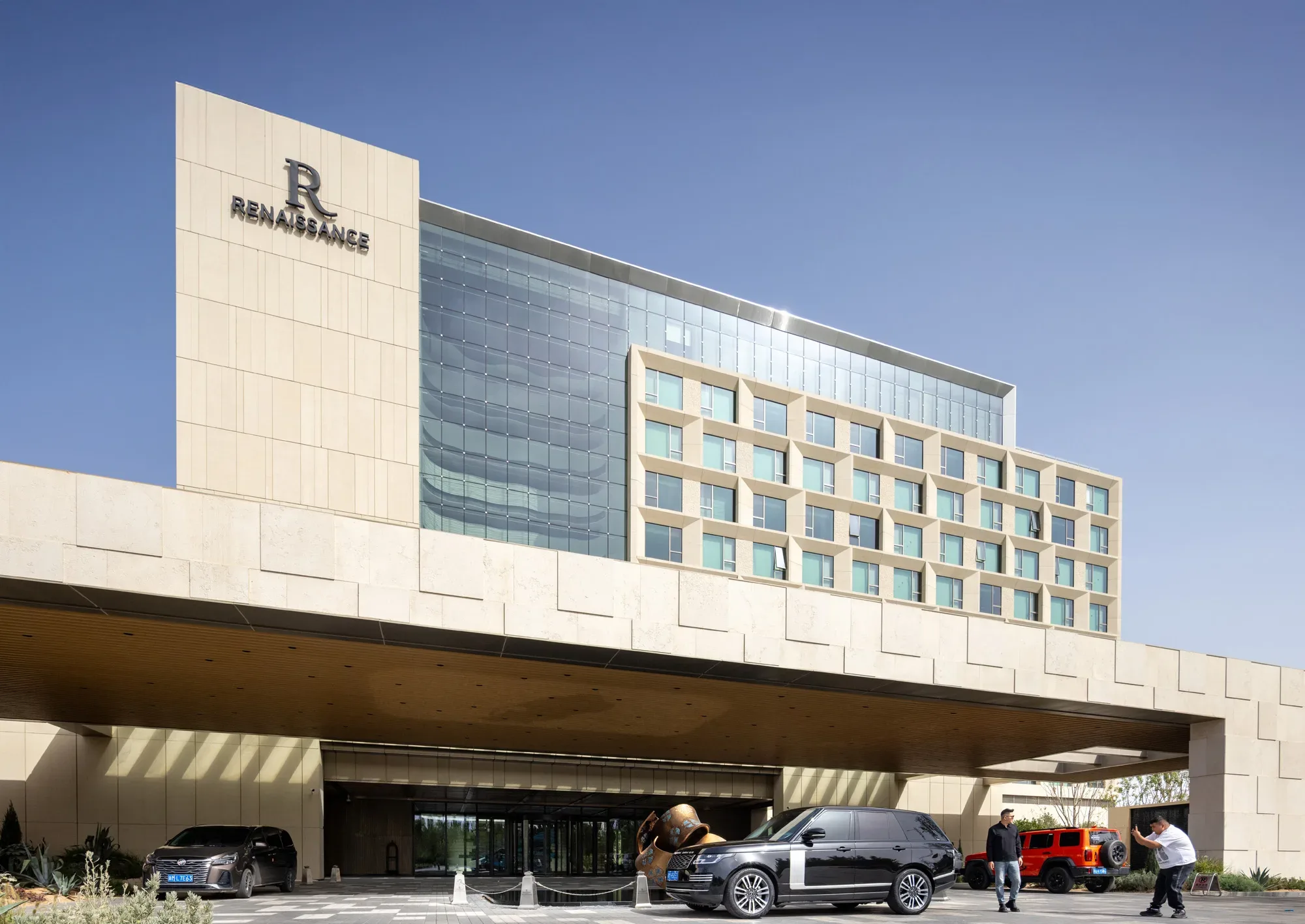 Aedas’ CGDG Korla dual‑brand hotel with layered stone façade, recessed glazing and sheltered drop‑off canopy. Street‑level view of the Aedas‑designed CGDG Korla Renaissance and Courtyard Hotels, showing a horizontally layered hotel mass with a stone‑clad façade and recessed floor‑to‑ceiling glazing. A deep cantilevered porte‑cochère defines the arrival sequence, while the upper levels feature repetitive window modules and a glazed corner volume marking shared hotel amenities.