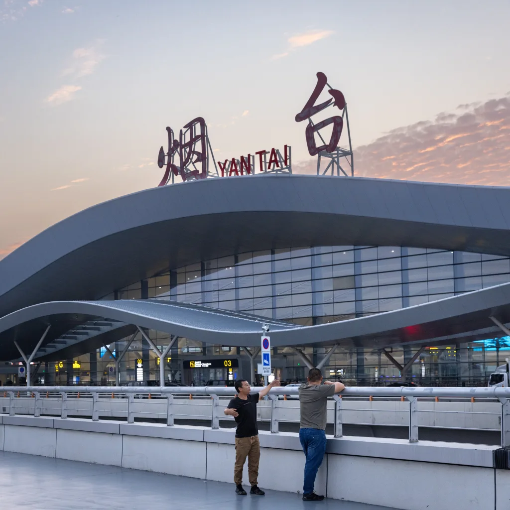 Aedas’ Yantai Penglai Airport Terminal 2 with wave‑form roof and glazed curtain wall reflecting an E‑shaped terminal layout. Dusk frontage view of Aedas’ Yantai Penglai International Airport Terminal 2 showing a continuous wave‑form roof over a unitised glass curtain wall. The terminal runs parallel to the runway, with a central glazed hall reflected in a water basin. Low‑profile landscaping and forecourt roads define the terminal’s landside interface in a coastal climate.