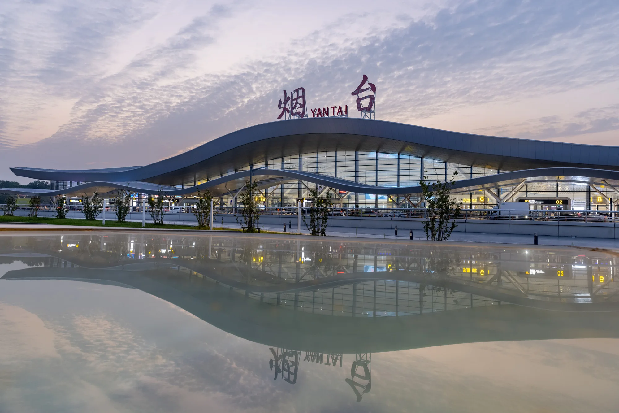 Aedas’ Yantai Penglai Airport Terminal 2 with wave‑form roof and glazed curtain wall reflecting an E‑shaped terminal layout. Dusk frontage view of Aedas’ Yantai Penglai International Airport Terminal 2 showing a continuous wave‑form roof over a unitised glass curtain wall. The terminal runs parallel to the runway, with a central glazed hall reflected in a water basin. Low‑profile landscaping and forecourt roads define the terminal’s landside interface in a coastal climate.