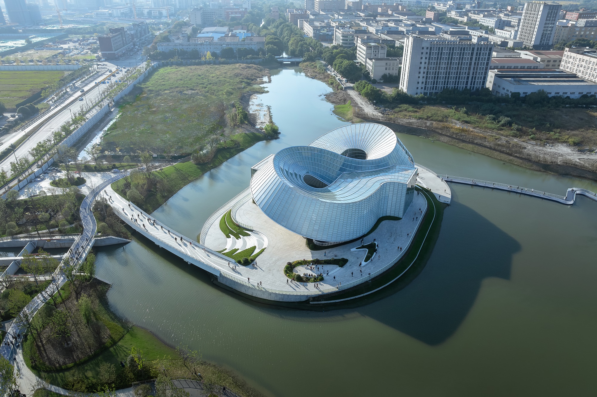Aedas’ Yohoo Museum, Hangzhou—double-ring glazed art museum on a lake island with pedestrian bridges. Aerial view of Aedas’ Yohoo Museum in Hangzhou: interlocking double-ring steel-frame volume wrapped in glazed curtain wall with vertical/horizontal framing. Two oculus courtyards pierce the undulating roof. Building sits on a lake island with promenade, planted terraces and pedestrian bridges—useful for daylighting strategy, flood-resilient waterfront design and parametric façade analysis.