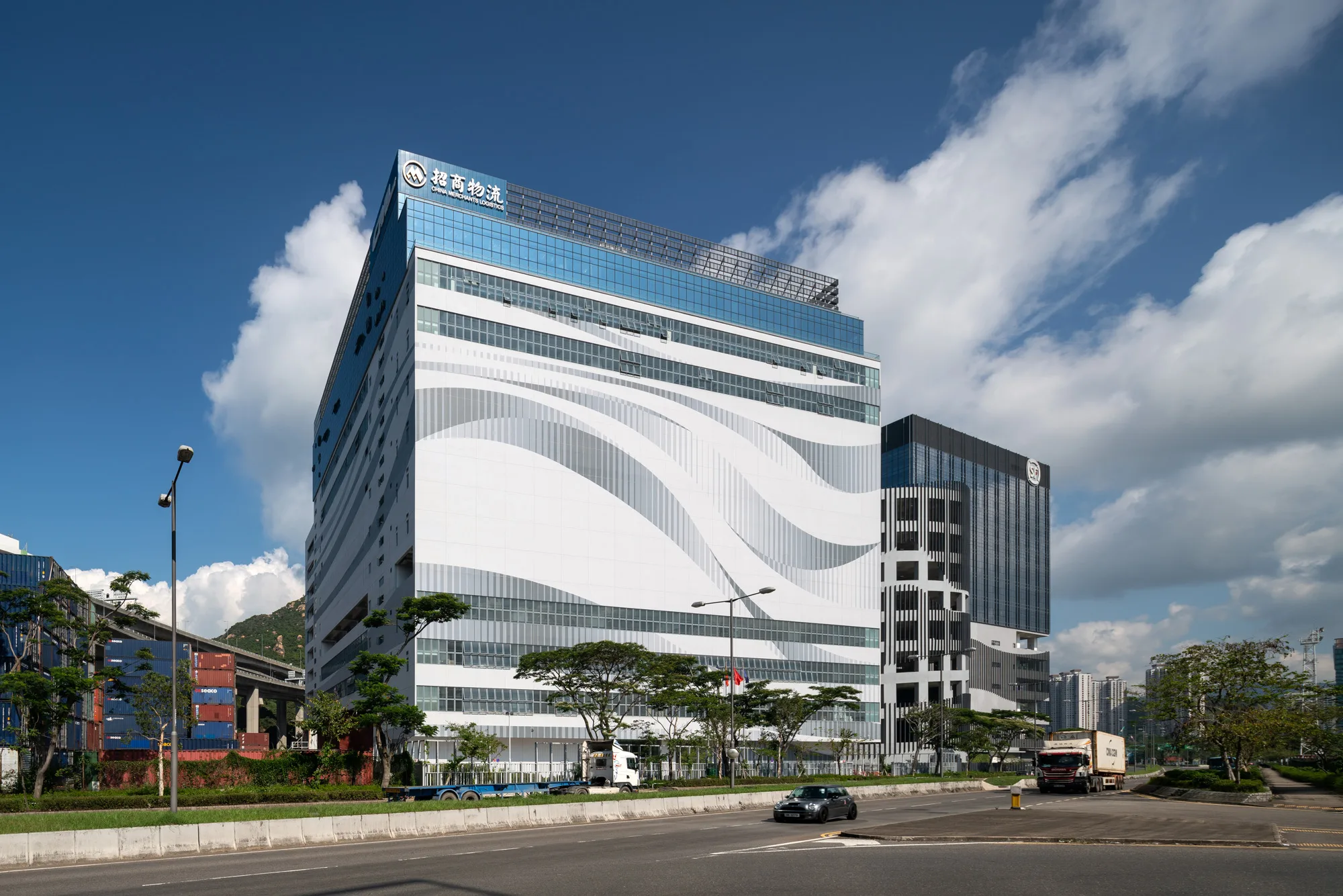 Aedas’ China Merchants Logistics Centre in Hong Kong is shown as a large‑scale logistics and office complex with a bold, wave‑patterned façade facing the waterfront. The image highlights a highly functional industrial building topped by cascading office terraces with extensive glazing, integrating efficient warehouse operations below with lighter, sea‑oriented office spaces above to create a distinctive logistics hub along the harbourfront.