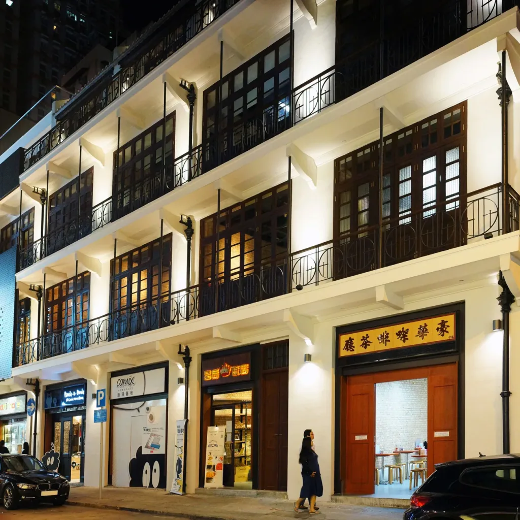 Aedas’ Revitalization of Mallory Street (Green House) in Hong Kong is shown as a restored row of pre‑war tenement buildings with cream‑coloured façades, timber shutters, wrought‑iron balconies, and illuminated ground‑floor shopfronts. The image highlights sensitive heritage conservation where original architectural details are preserved and reactivated, creating a vibrant street‑level cultural destination that blends historical character with contemporary creative uses in Wan Chai’s dense urban fabric.