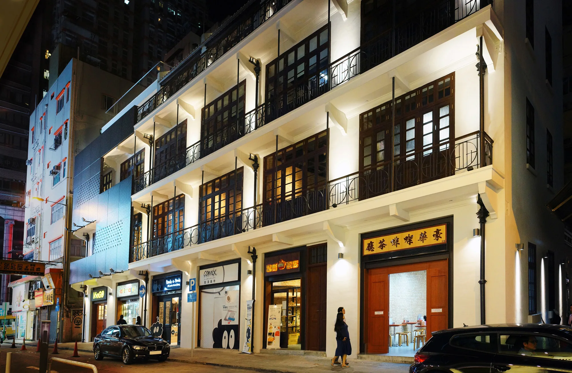 Aedas’ Revitalization of Mallory Street (Green House) in Hong Kong is shown as a restored row of pre‑war tenement buildings with cream‑coloured façades, timber shutters, wrought‑iron balconies, and illuminated ground‑floor shopfronts. The image highlights sensitive heritage conservation where original architectural details are preserved and reactivated, creating a vibrant street‑level cultural destination that blends historical character with contemporary creative uses in Wan Chai’s dense urban fabric.
