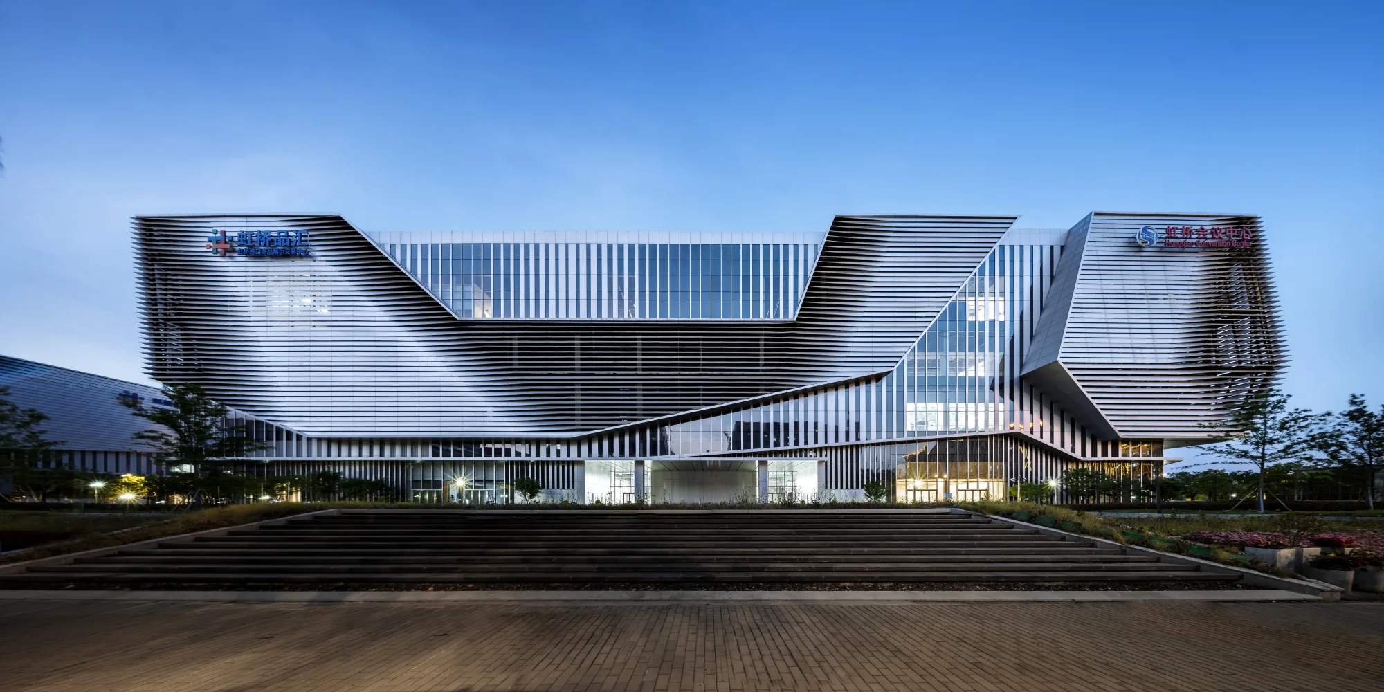 Aedas’ Shanghai Hongqiao Import Commodity Exhibition and Trade Center is shown as a low‑rise, sculptural commercial complex with dramatic cantilevered horizontal louvers and deep, folded façades framing a central glazed volume. The image highlights a bold, contemporary architectural expression that emphasises depth and movement, supporting a hybrid exhibition‑retail environment with strong visual presence and clear urban connectivity near Hongqiao Airport.