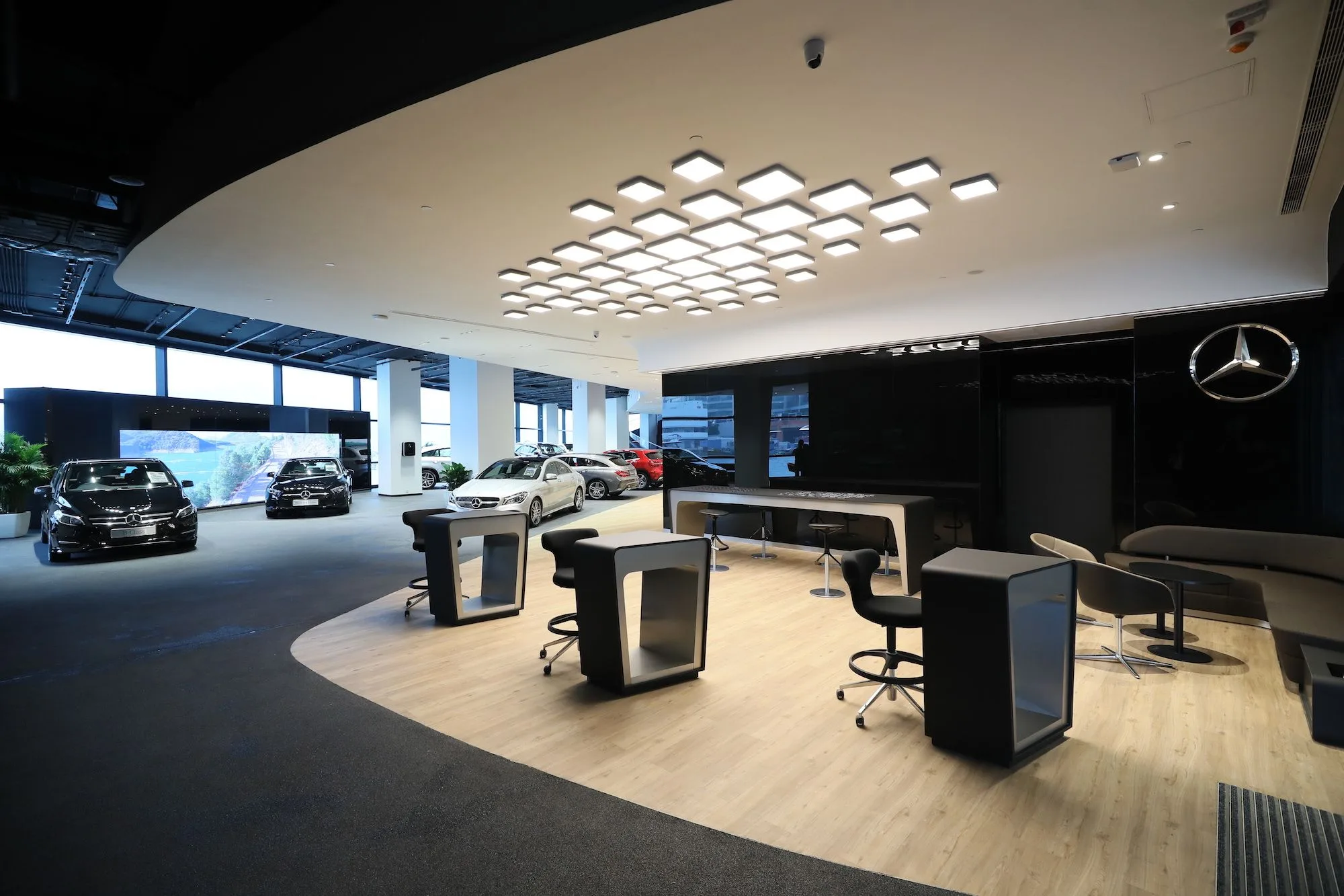 Aedas’ Mercedes Showroom interior in Hong Kong is shown as a sleek, open‑plan automotive gallery featuring a curved illuminated ceiling, polished light‑wood flooring, and minimalist consultation desks. The image highlights a refined showroom environment where premium vehicles are displayed within a calm, modern setting, integrating brand identity, customer interaction zones, and expansive glazing to support an immersive automotive sales experience.