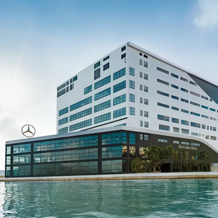Aedas’ Mercedes Showroom in Hong Kong is shown as a multi‑storey automotive branding center with a crisp white upper volume set above a transparent, glass‑wrapped showroom podium along the waterfront. The image highlights a bold conversion of an industrial building into a contemporary Mercedes‑Benz flagship, featuring expansive display glazing, integrated signage, and a strong horizontal base that connects showroom, service facilities, and rooftop event space into a unified harbourfront landmark.