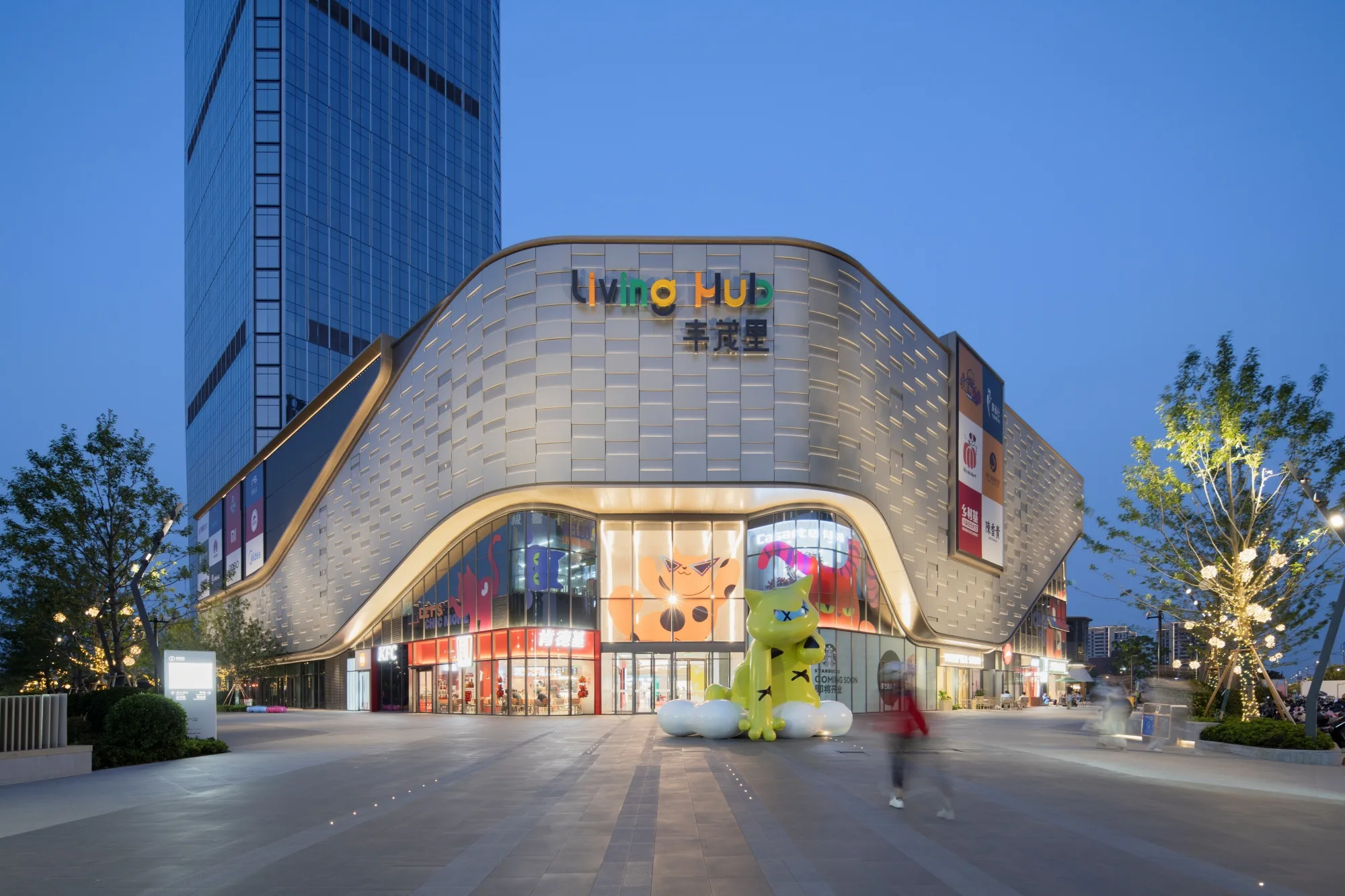 Aedas’s Changshu Jinmao Living Hub is shown as a mixed‑use urban complex featuring a sculpted retail podium with curved metallic façade panels and an integrated high‑rise tower. The street‑level view highlights inward‑focused commercial circulation, active plaza frontage, and contemporary façade articulation within Xuzhou’s regional commercial context.