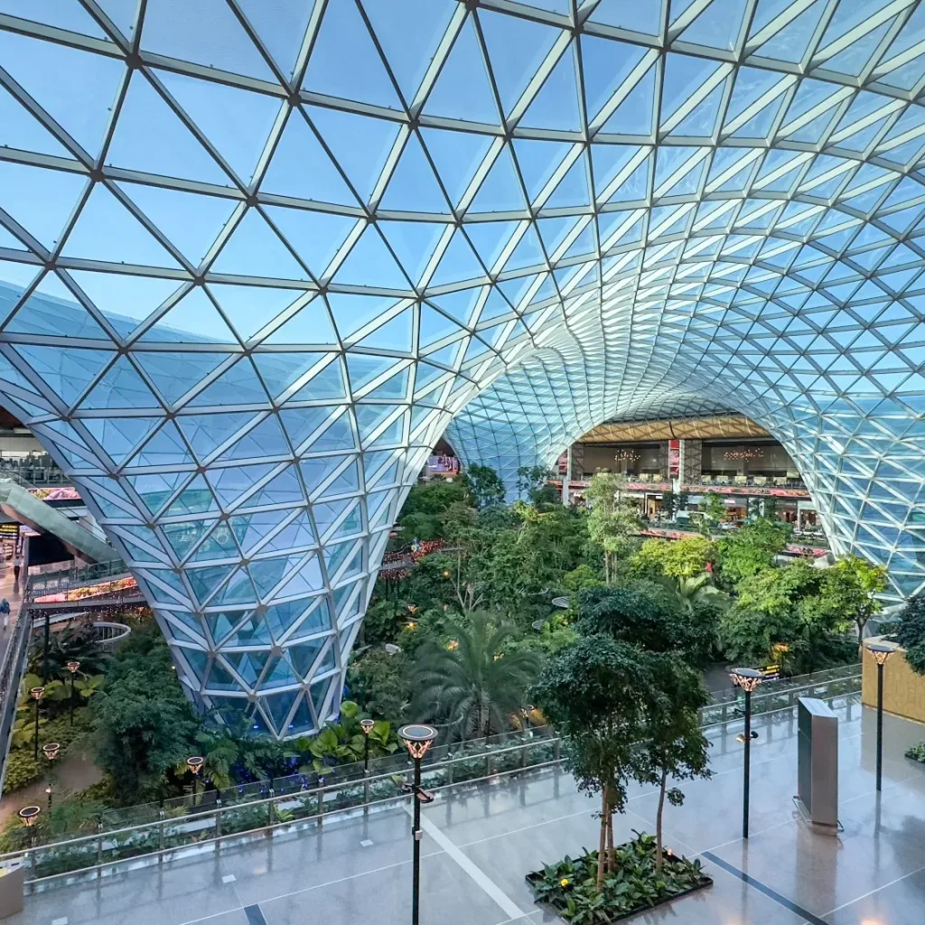 Aedas’ HIA Central Concourse with column‑free grid shell roof over indoor tropical garden beneath glazed skylights, Doha. Interior view of the Aedas-designed Central Concourse at Hamad International Airport, Doha, showing an 85‑metre column‑free steel grid shell roof with triangulated glazing. Natural light filters through the roof structure onto a large indoor tropical garden with trees, water features and elevated passenger walkways, integrated within the airport’s climate‑controlled terminal environment.