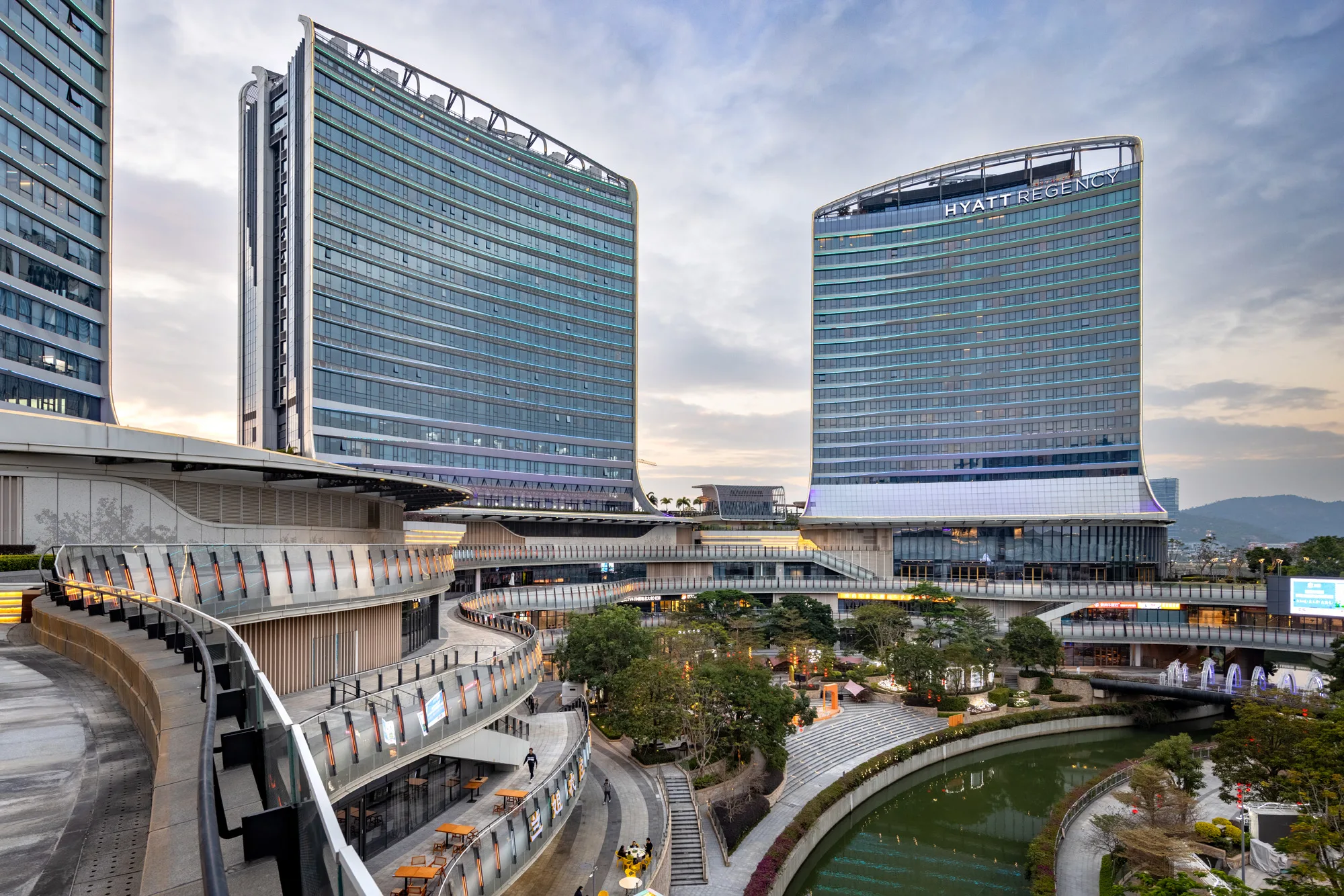 Aedas’ Novotown mixed‑use destination with terraced retail decks, twin hotel towers and landscaped water plaza in Hengqin. Oblique view of Aedas’ Novotown showing two glazed hotel towers rising from a multi‑level podium. Curved, terraced retail decks step down toward a central landscaped water feature. The façades use unitised glass curtain walls and horizontal slab edges, integrating pedestrian circulation, public plazas and entertainment uses within a subtropical urban context.