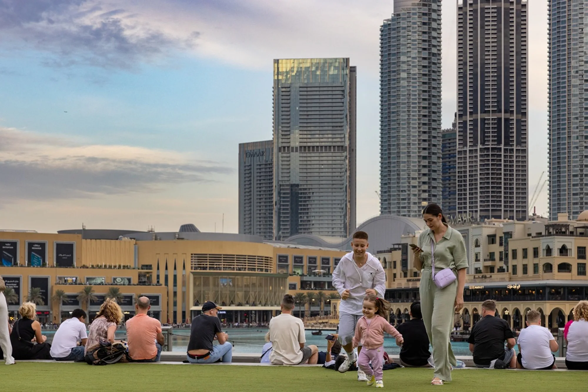 Aedas’ Vida Dubai Mall twin towers above a stepped podium, framing a public lawn in Downtown Dubai. Twilight view of a public lawn in Downtown Dubai facing Aedas‑designed Vida Dubai Mall. Two slender high‑rise towers rise behind a stepped podium edge, with tower façades expressed by vertical glazing bands. The foreground shows an active public realm with seated visitors and open sightlines toward the skyline.