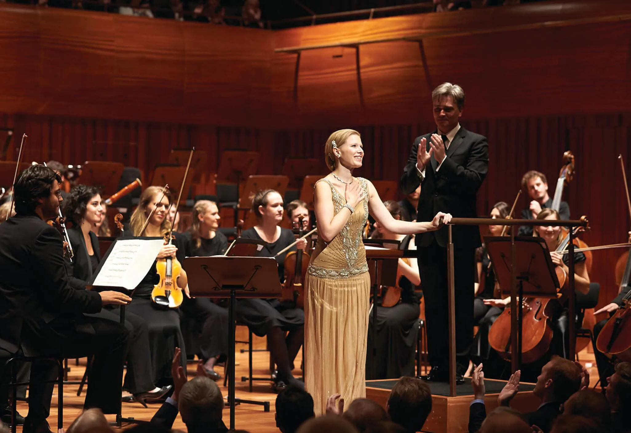 Aedas’s Milton Court concert hall at the Guildhall School of Music & Drama is shown with a central performance stage, raked audience seating, curved timber acoustic wall panels, and integrated stage lighting. The image highlights acoustic‑driven interior architecture, performance‑optimized sightlines, and large‑capacity cultural venue design in an urban London context.