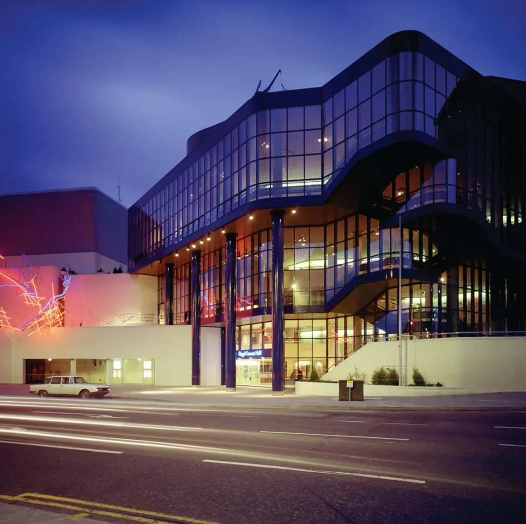 Aedas’s Nottingham Royal Concert Hall is shown as a large civic cultural building with a multi‑storey glazed façade, curved upper volumes, and a solid plinth base. The night‑time exterior highlights performance‑led architecture, transparent public interfaces, and integration with the urban streetscape in a UK city centre context.