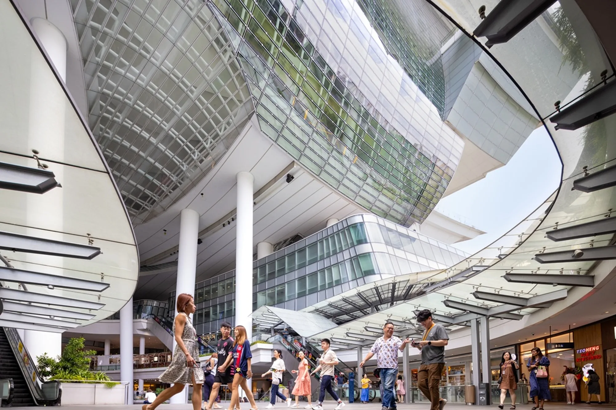 Aedas’ The Star in Singapore with elevated auditorium, open‑air retail podium and climate‑responsive structural canopy. Interior plaza view of Aedas‑designed The Star in Singapore, showing a vast open‑air atrium beneath an elevated auditorium supported by inclined concrete columns. Retail floors, escalators and staircases wrap the central void, while a high‑performance glazed façade and overhead roof structure modulate daylight and airflow within the public space.