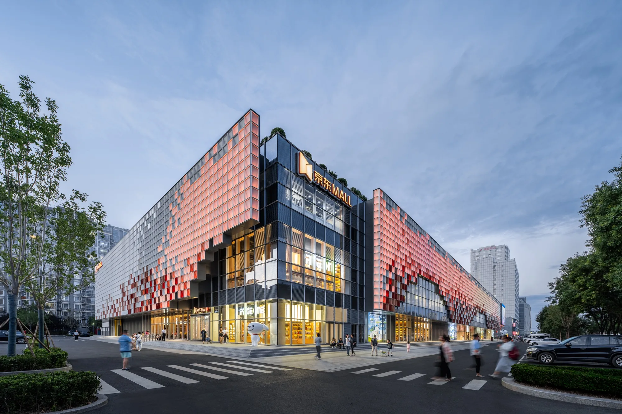 Aedas’ Beijing JD Mall Renovation is shown as a revitalised urban retail corner with a bold pixelated façade composed of layered red and silver panels wrapping a glazed volume. The image highlights a contemporary architectural expression where light, shadow, and a grid‑based skin reinterpret digital pixels and traditional brick textures, creating a dynamic, technology‑driven landmark that activates the streetscape with transparent entrances and renewed public engagement.