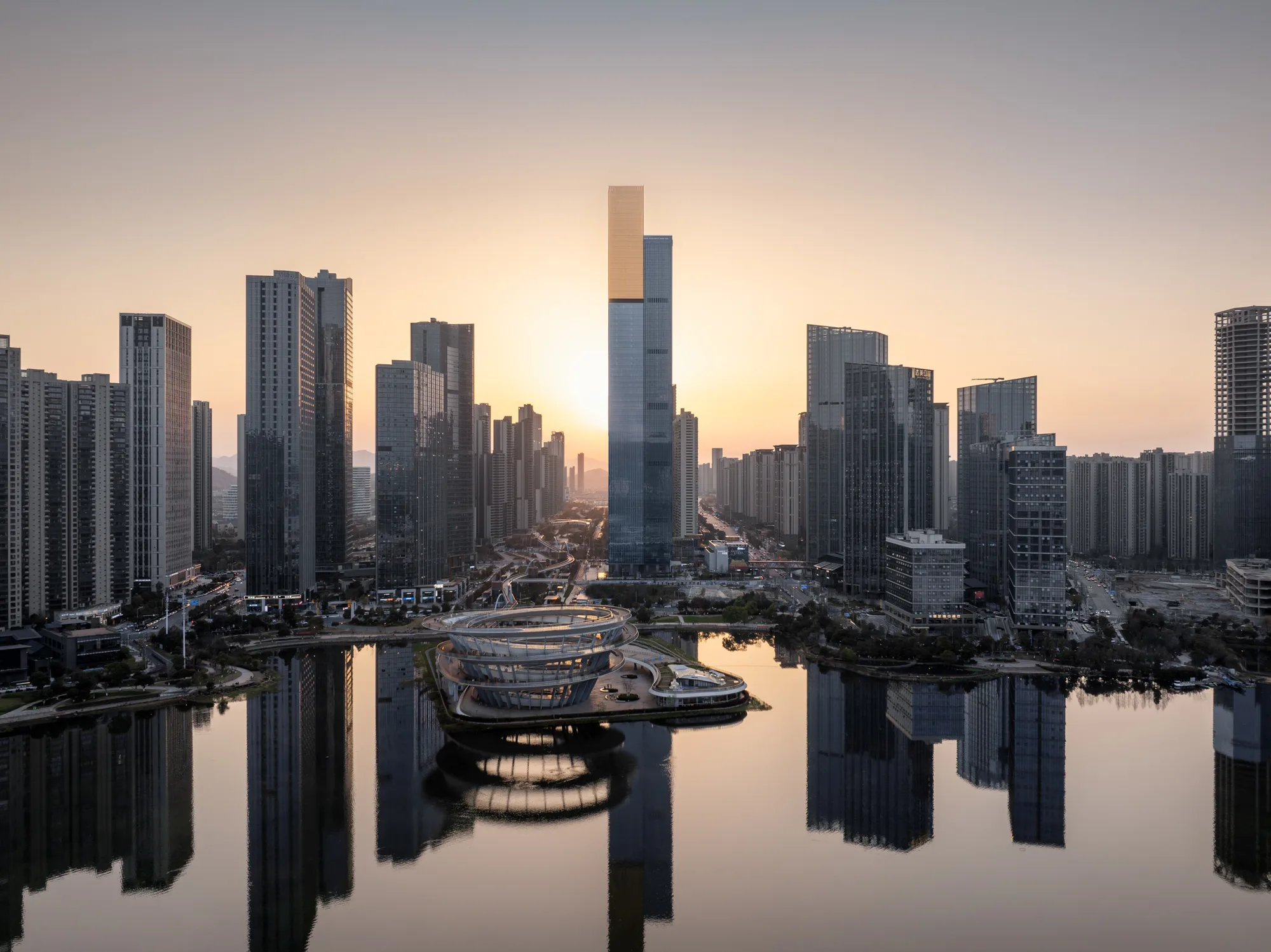 Aedas’ Jinmao Tower - 318 m mixed‑use skyscraper in Changsha with stepped podium, lakefront setting and high‑performance glazed tower. Aerial dusk view of an Aedas-designed mixed-use development in Changsha featuring a slender supertall tower rising above a stepped podium and central courtyard. The tower uses high-performance curtain wall glazing, fronting a lake and dense high-rise city blocks at a major urban intersection near metro infrastructure.